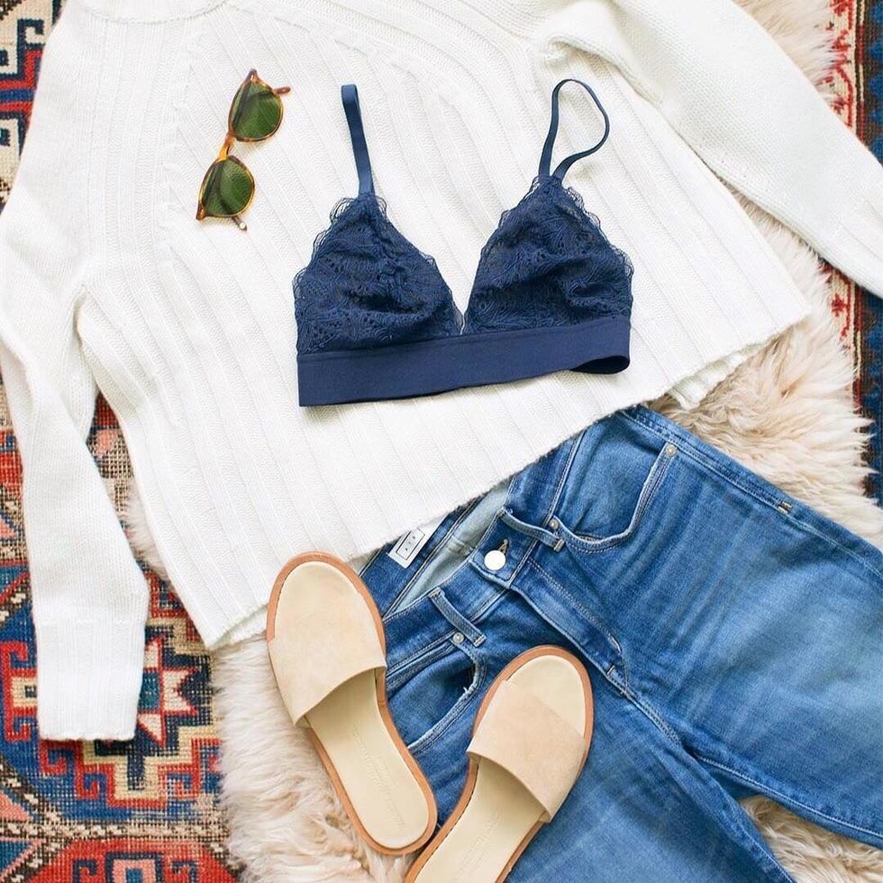 A Lively navy blue lace bralette lays on a white ribbed fall sweater with a pair of blue jeans, nude slide sandals, and sunglasses laying on an Aztec rug